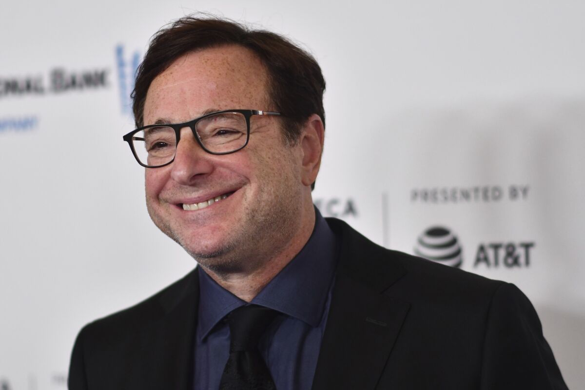 Bob Saget, in a black suit and tie, smiles for cameras at a premiere