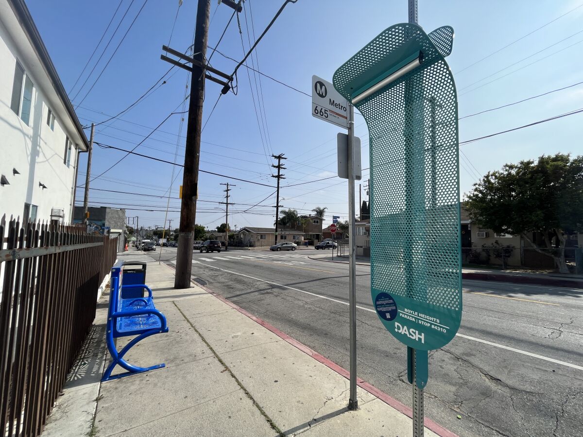 A perforated green metal screen shaped like a skateboard is attached to bus stop pole on a sunny L.A. street