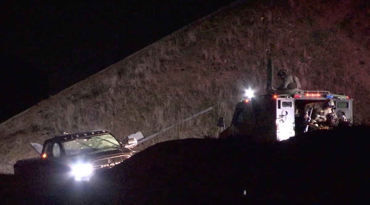 Officers in an armored vehicle point guns and lights at a Ram pickup in a ditch with several bullet holes in its windshield