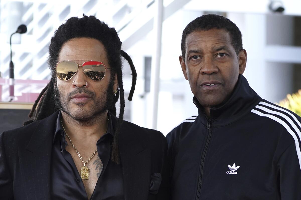 Lenny Kravitz in mirrored sunglasses standing next to Denzel Washington, clad in a black tracksuit jacket with white stripes