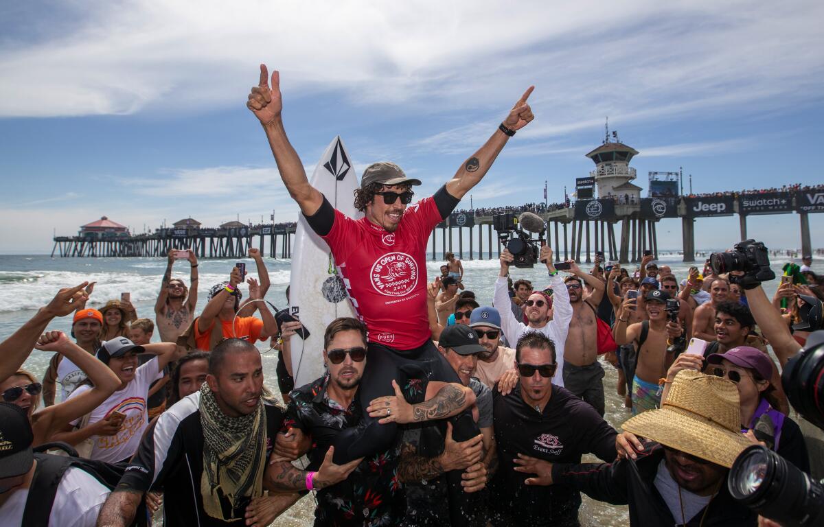 Amid a crowd, a man is held aloft, raising both arms. Others take photos. In the background are the ocean and a pier.