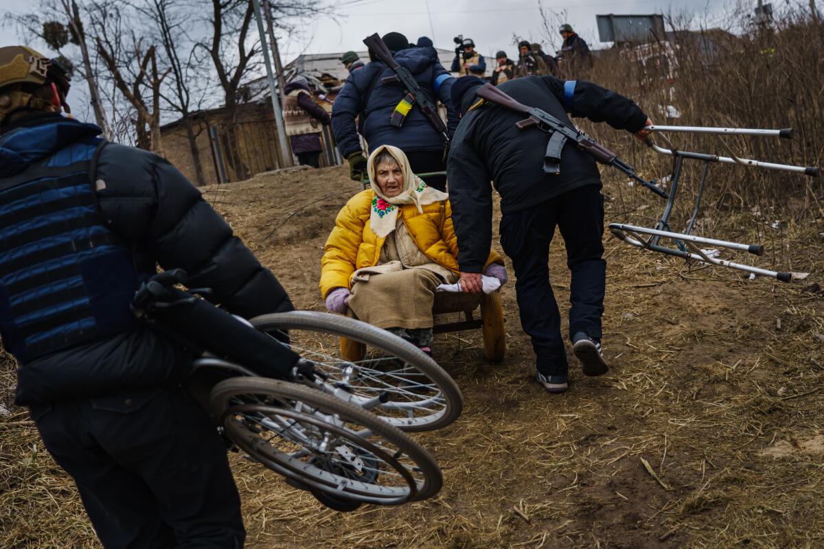 an old woman in a yellow coat sits in a wheelbarrow as soldiers carry her up a hill