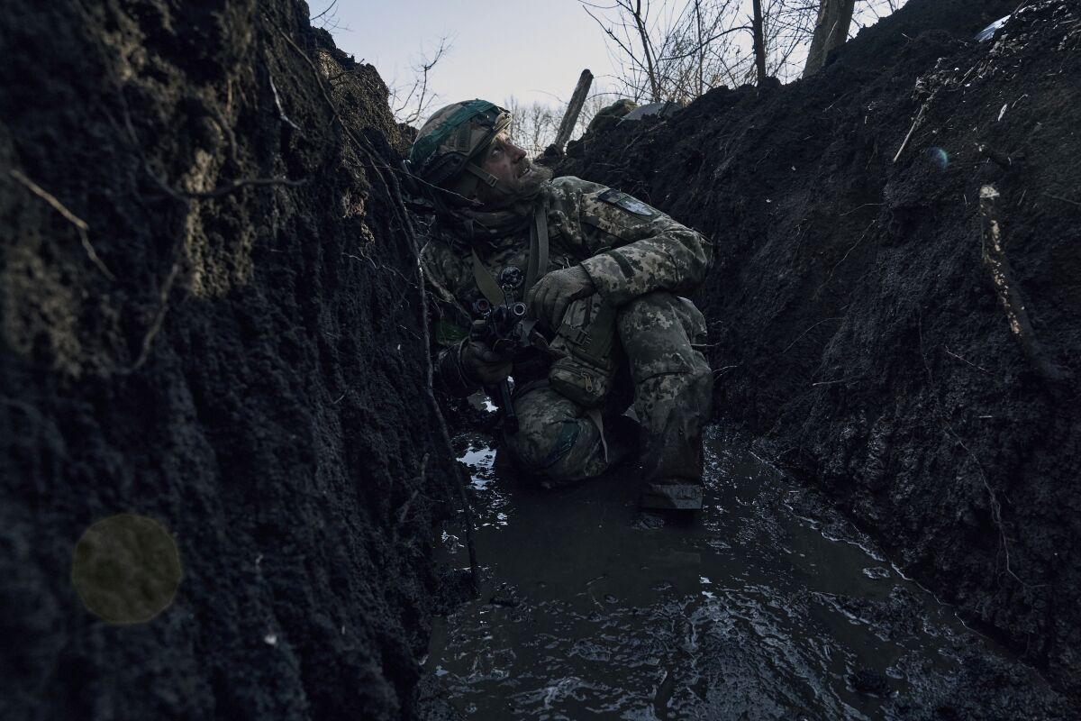 A man in military fatigues looks skyward while crouched in a muddy trench