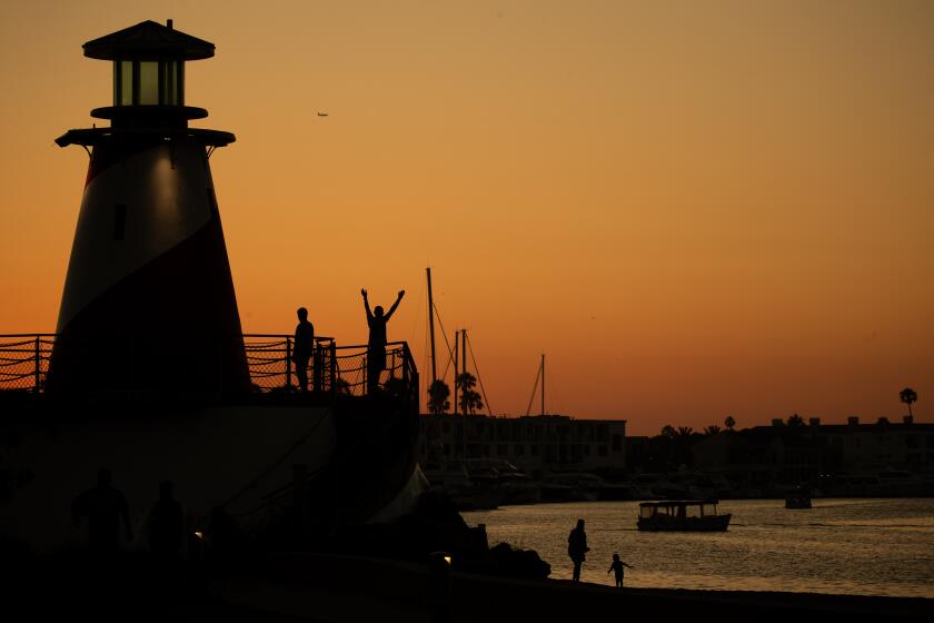 Newport Beach, CA - July 19: Amid a golden sky, a couple look out from a playground lighthouse while an adult and child explore the water's edge on a warm summer evening at dusk at Marina Park, Newport Harbor in Newport Beach Wednesday, July 19, 2023. (Allen J. Schaben / Los Angeles Times)