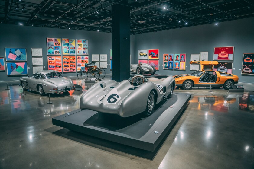 Walls lined with paintings by Andy Warhol and several cars inside a gallery.