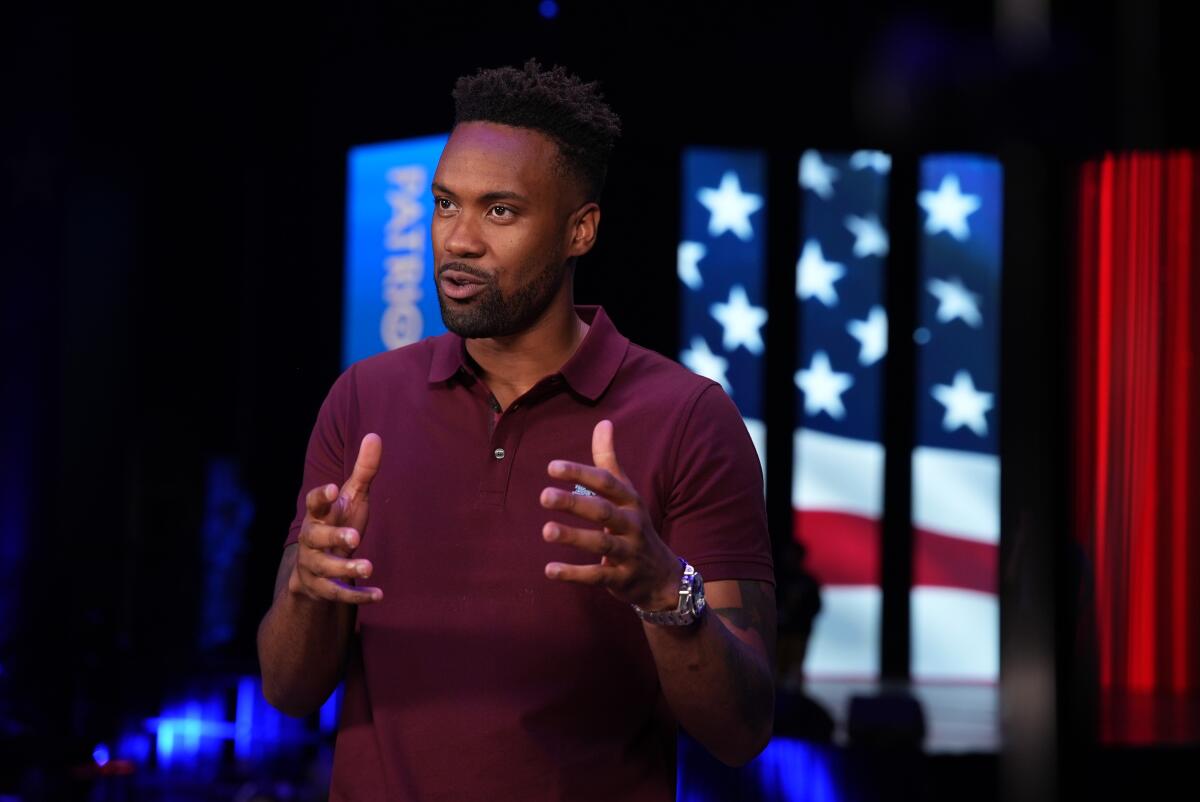 Lawrence Jones in a burgundy polo shirt gestures with his hands seated in front of a cutout of an American flag.