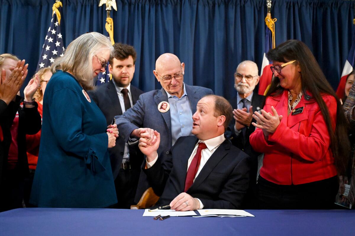 Seated behind a desk, Gov. Jared Polis hands his pen to Sandy and Lonnie Phillips.