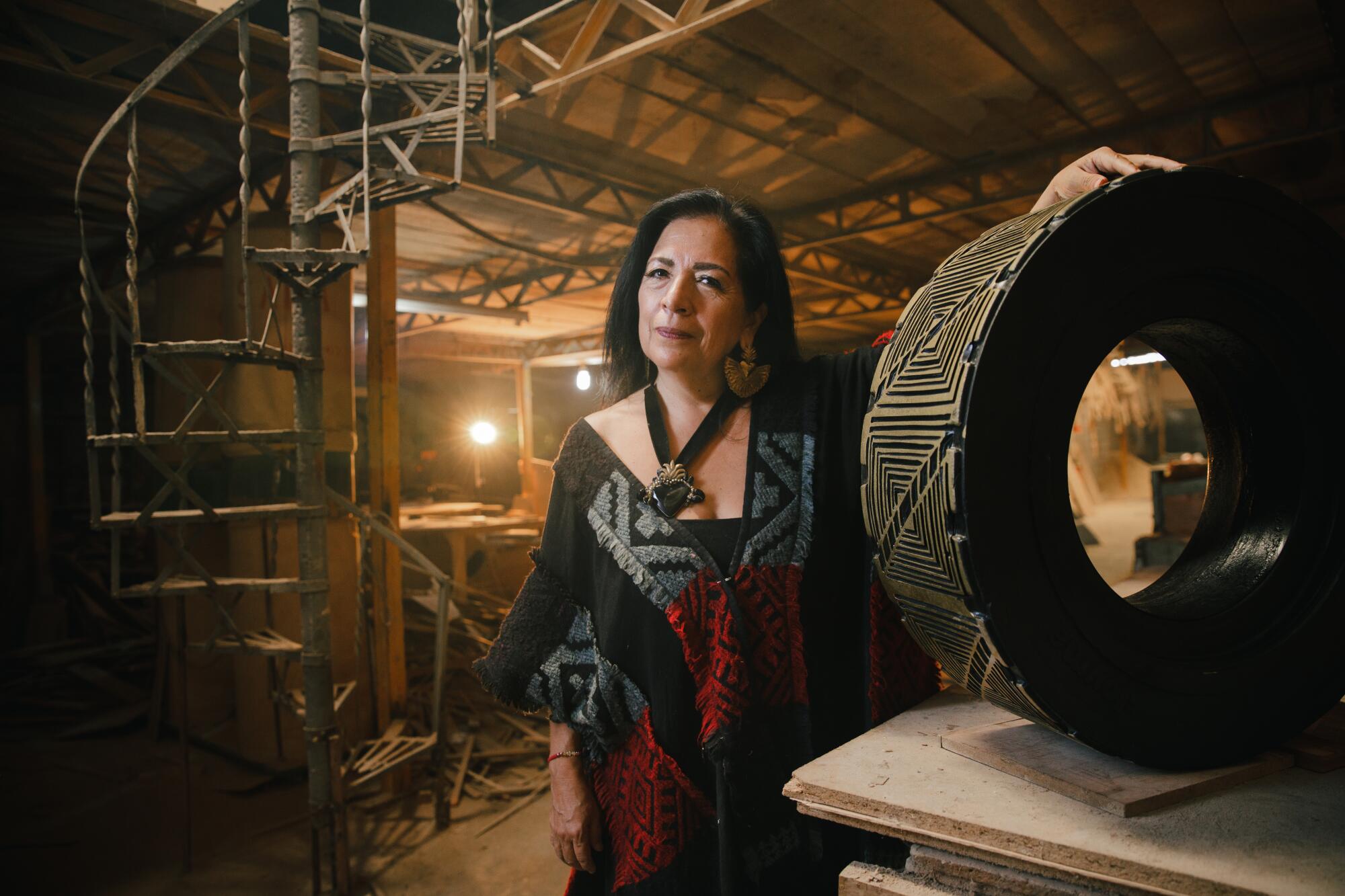 With an industrial studio as backdrop, a woman stands next to a vehicle tire carved and painted into  sculpture. 