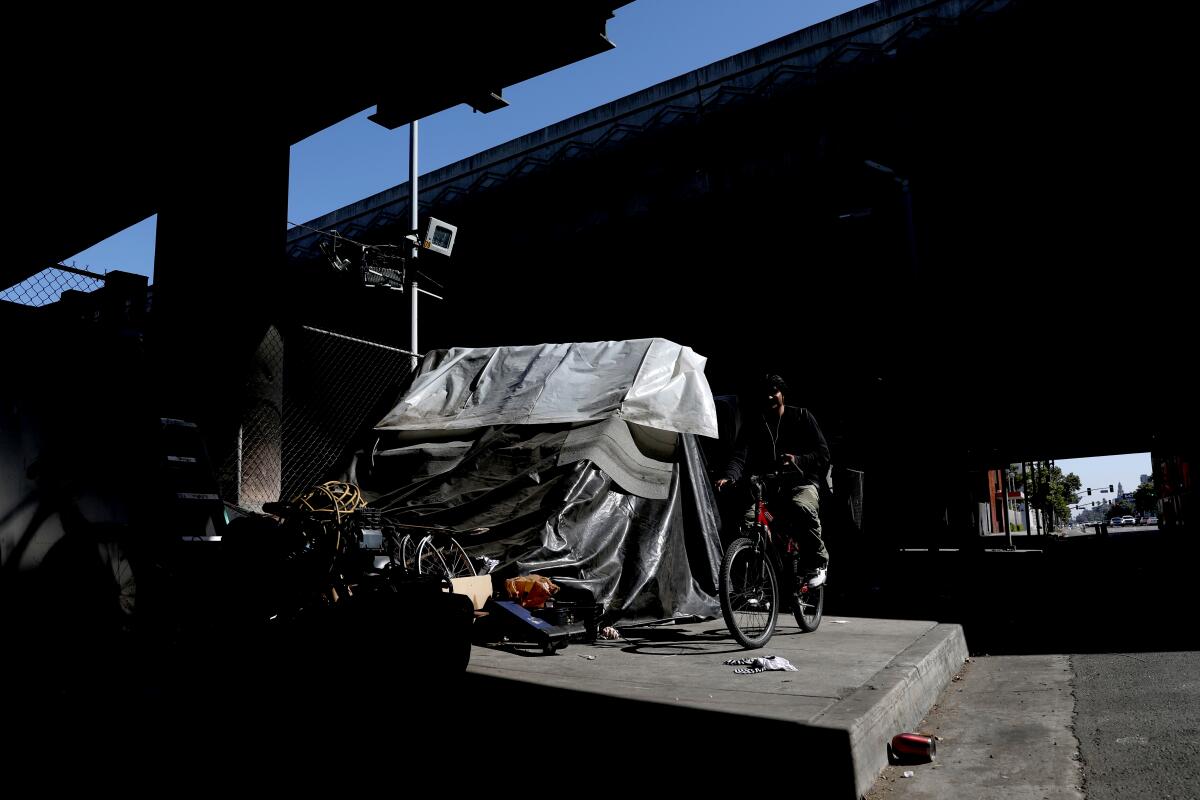 A person rides past a homeless encampment on the sidewalk underneath an overpass 