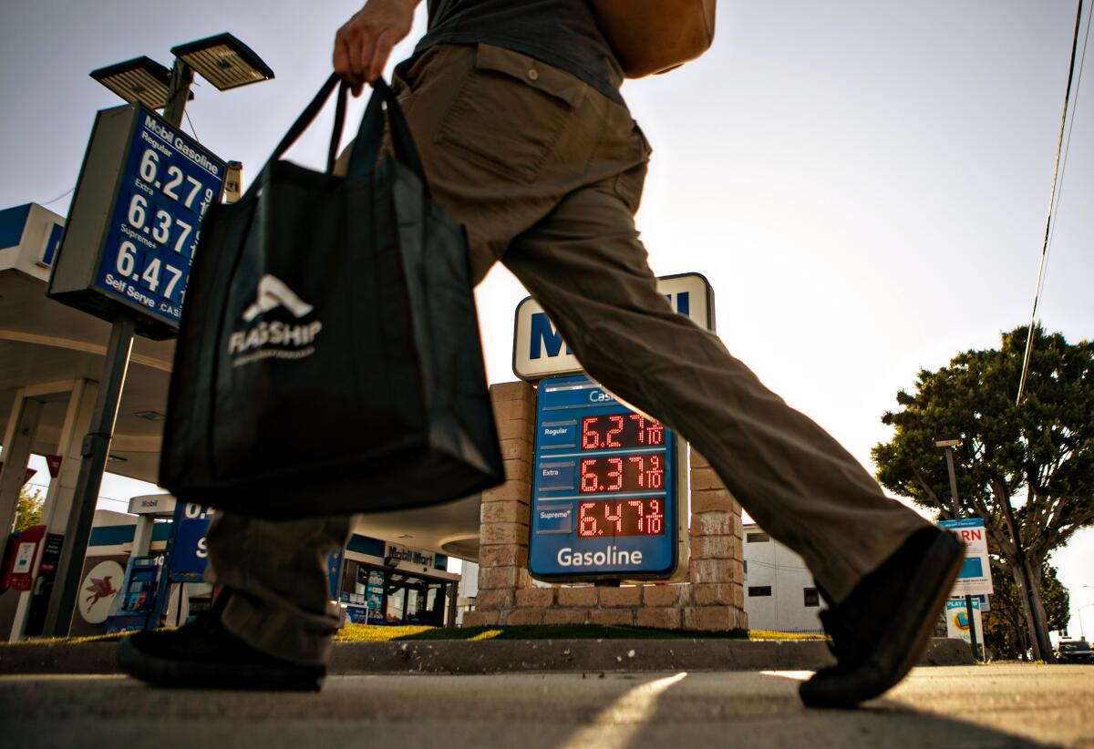A man carrying a bag walks past a couple of signs with prices outside a gas station.