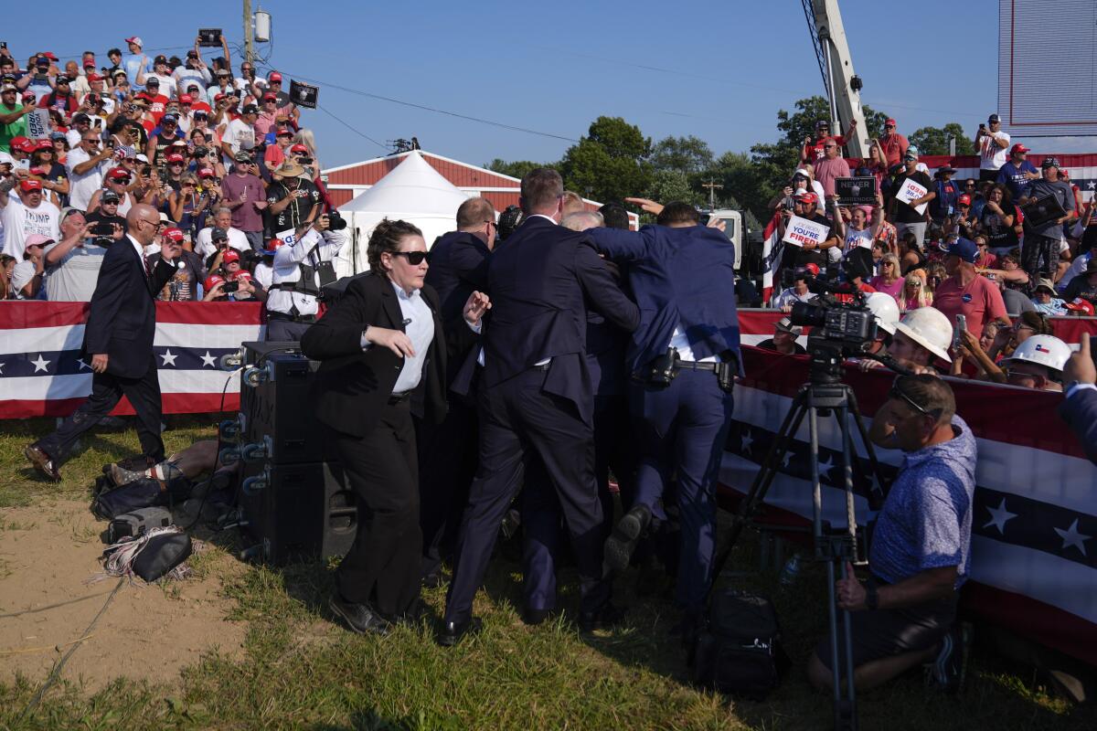 Suited security officers surround former President Trump as spectators watch from stands draped in the colors of the American flag.