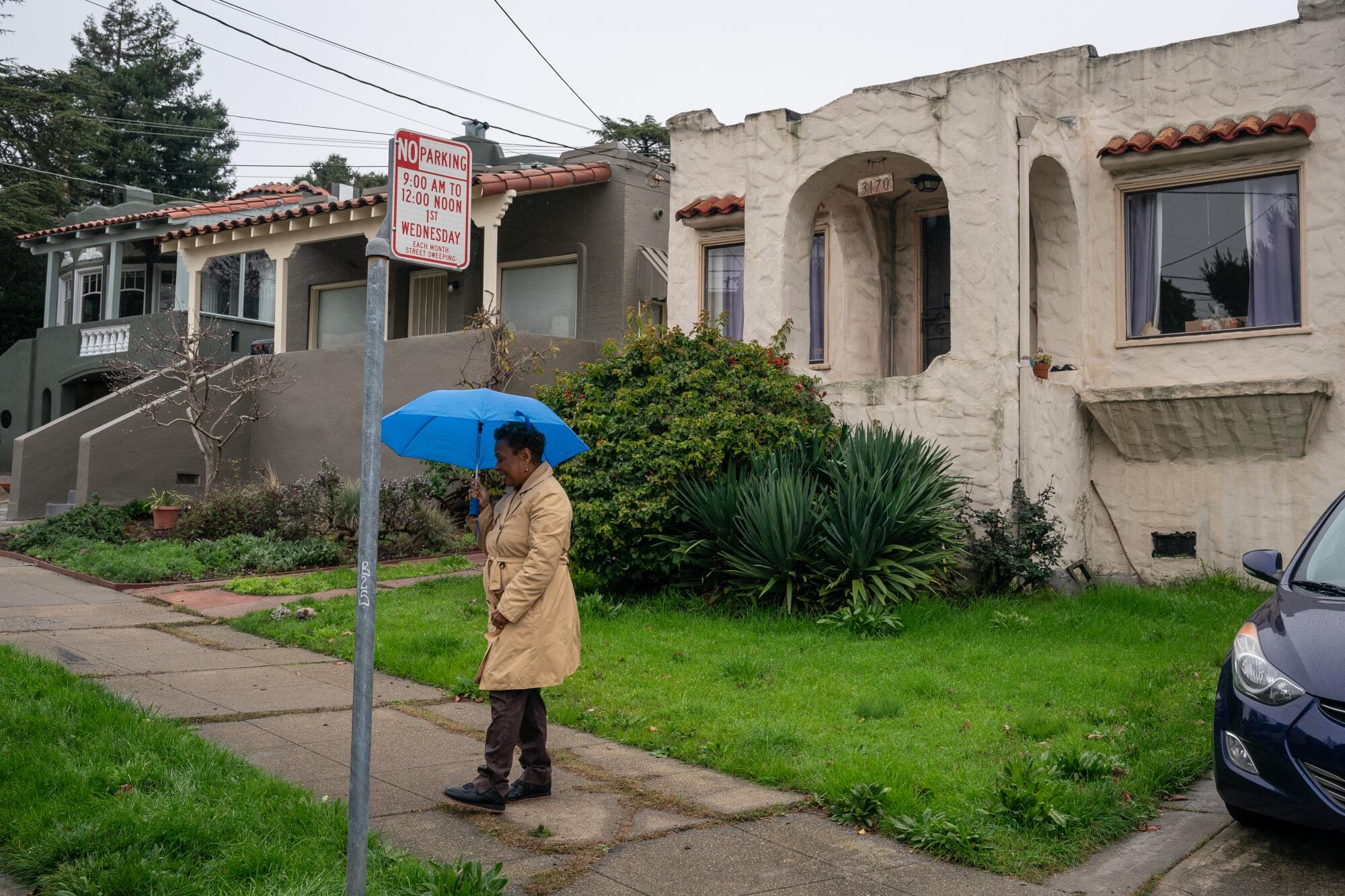 Barbara Lee stands on a sidewalk with an umbrella and in a coat in front of a house. 