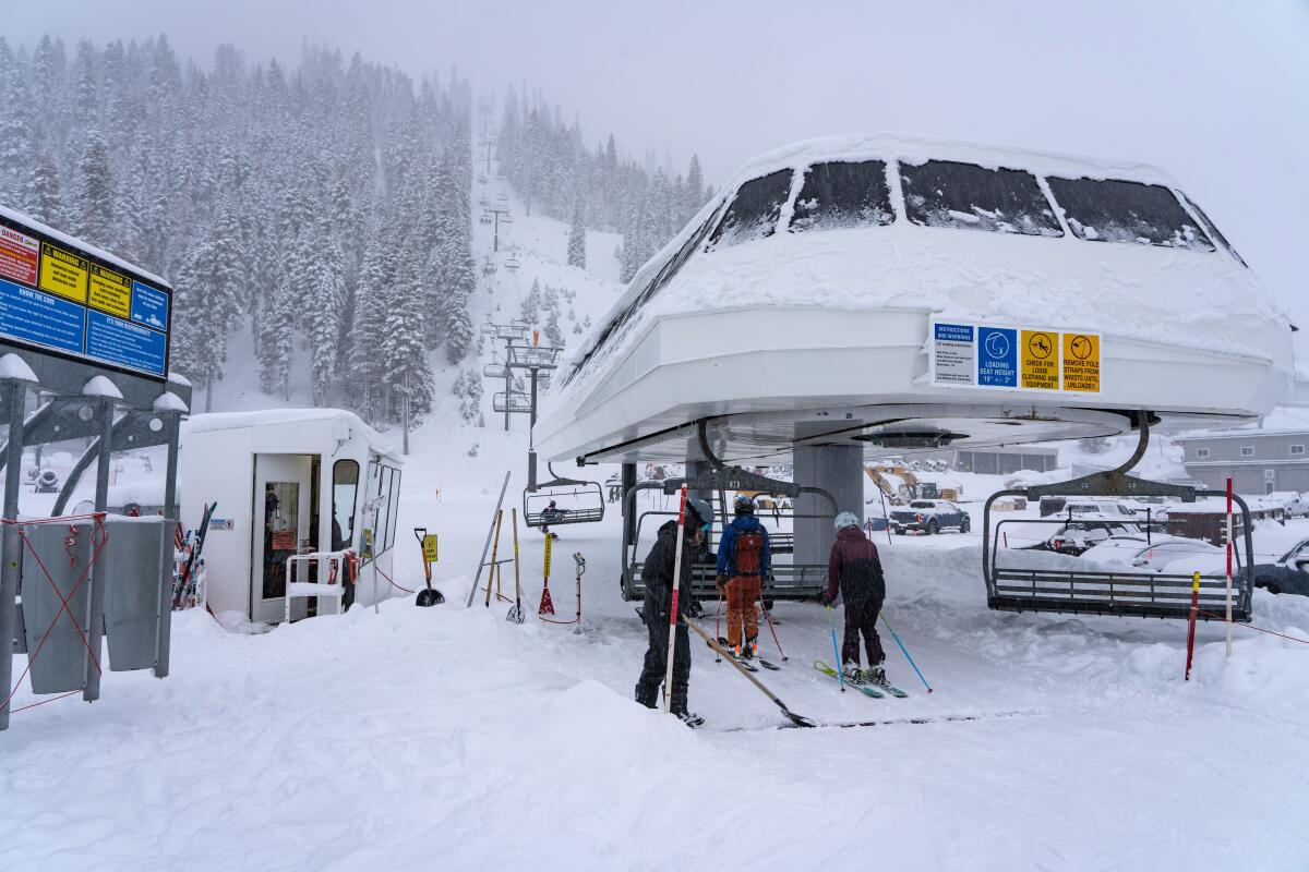 People on skis prepare to sit down on a lift at the bottom of a snowy mountain.