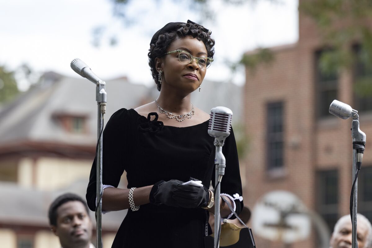 A Black woman in 1960s mourning attire stands at a microphone in a scene from a movie