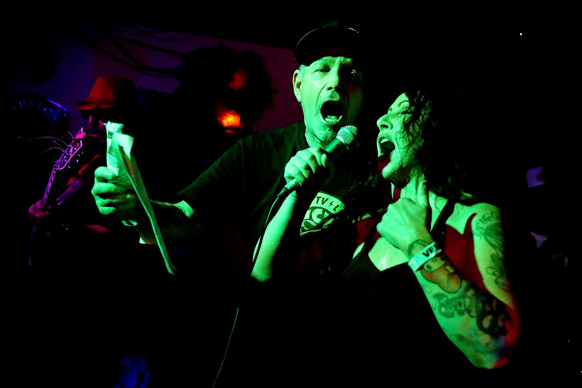 Two karaoke singers belt out a song in a dark lit room, under lights that make their skin appear to be green.