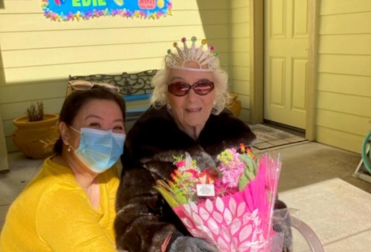 Edie Ceccarelli, right, wearing a tiara and holding flowers, with her caregiver kneeling beside her.