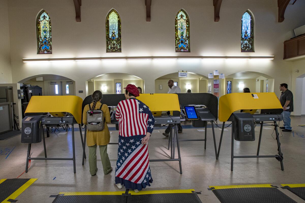 A woman votes in a modern voting booth. Behind her stands a woman in a flag-adorned jacket and skirt. 

