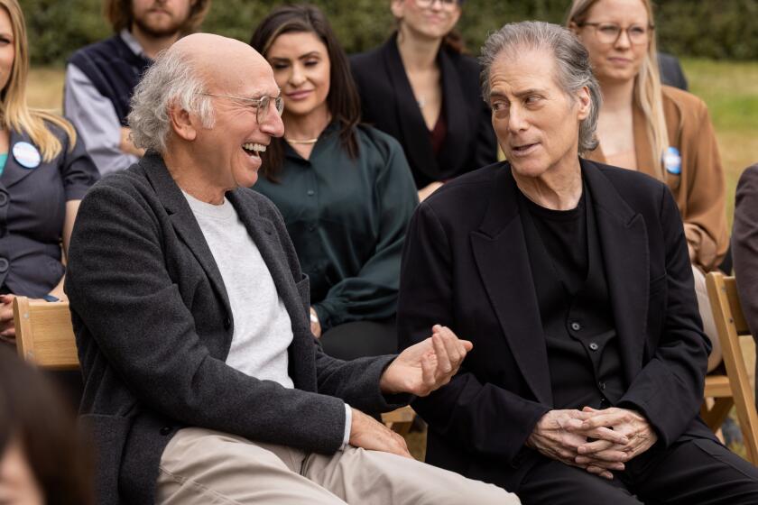 Larry David in a grey shirt and black suit jacket smiling and sitting next to Richard Lewis in a dark outfit