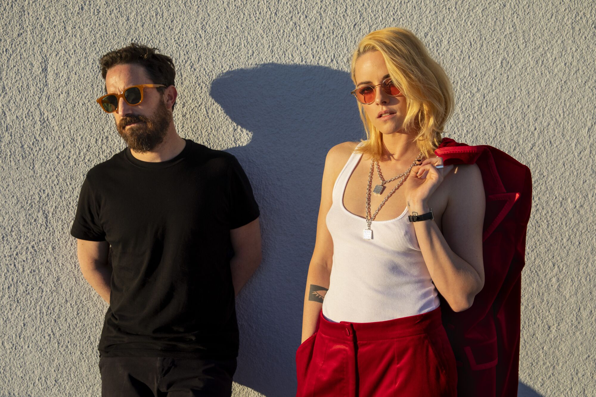 A man in black and a woman in a white tank top and red pants, both wearing sunglasses, lean against a wall on a sunny day.