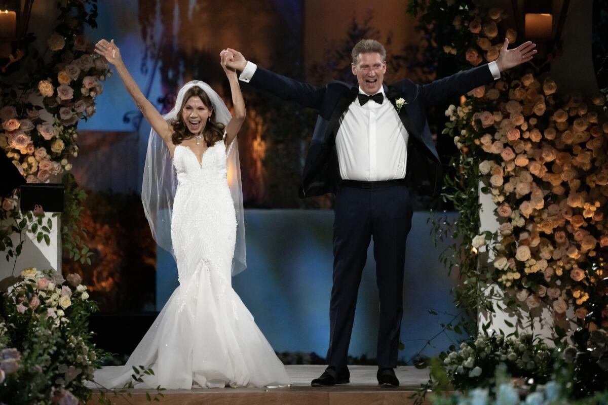 A smiling woman in a white wedding dress and a man in a tuxedo raise their arms in the air.