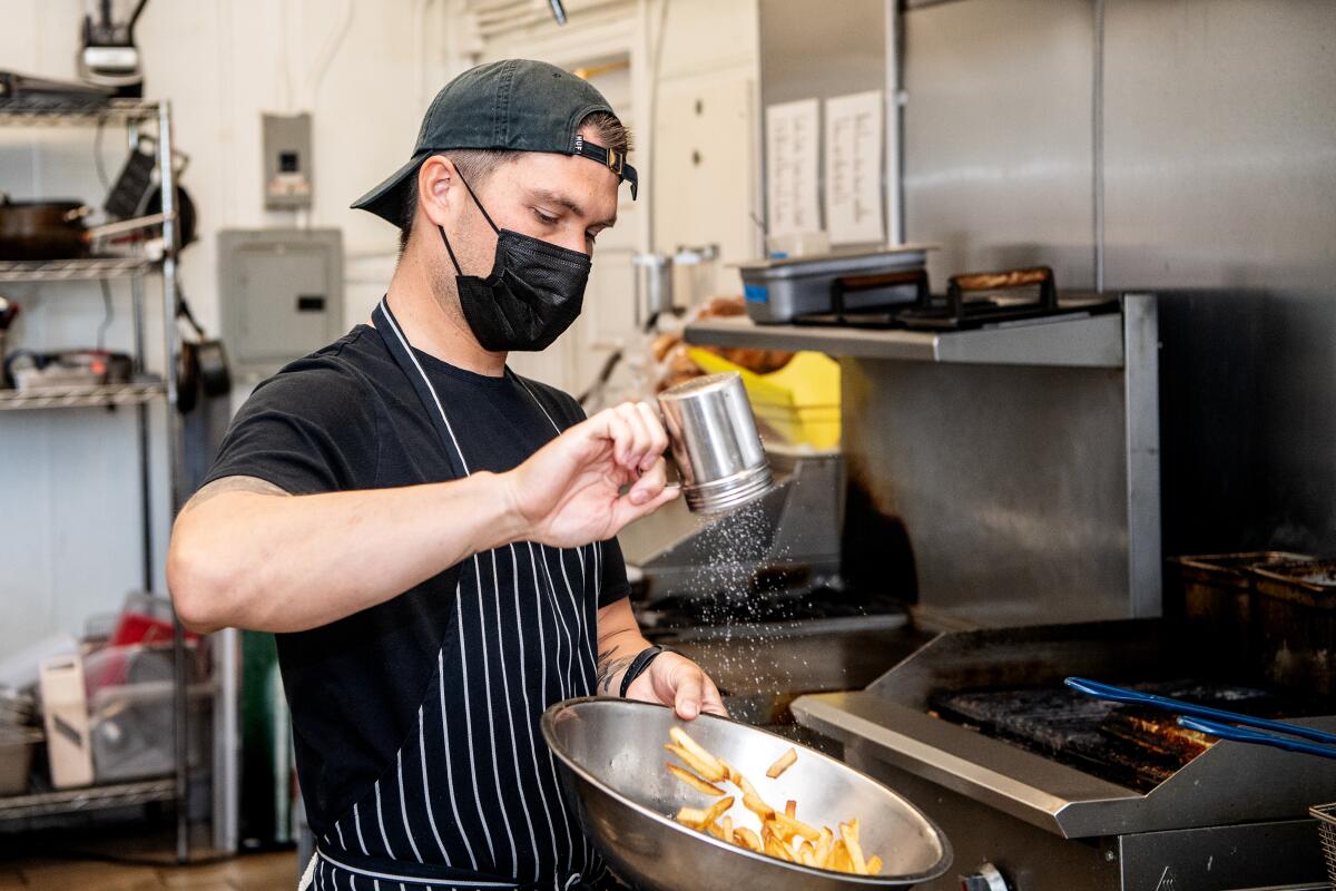 A man in a mask sprinkles salt into a large bowl of fries in a commercial kitchen
