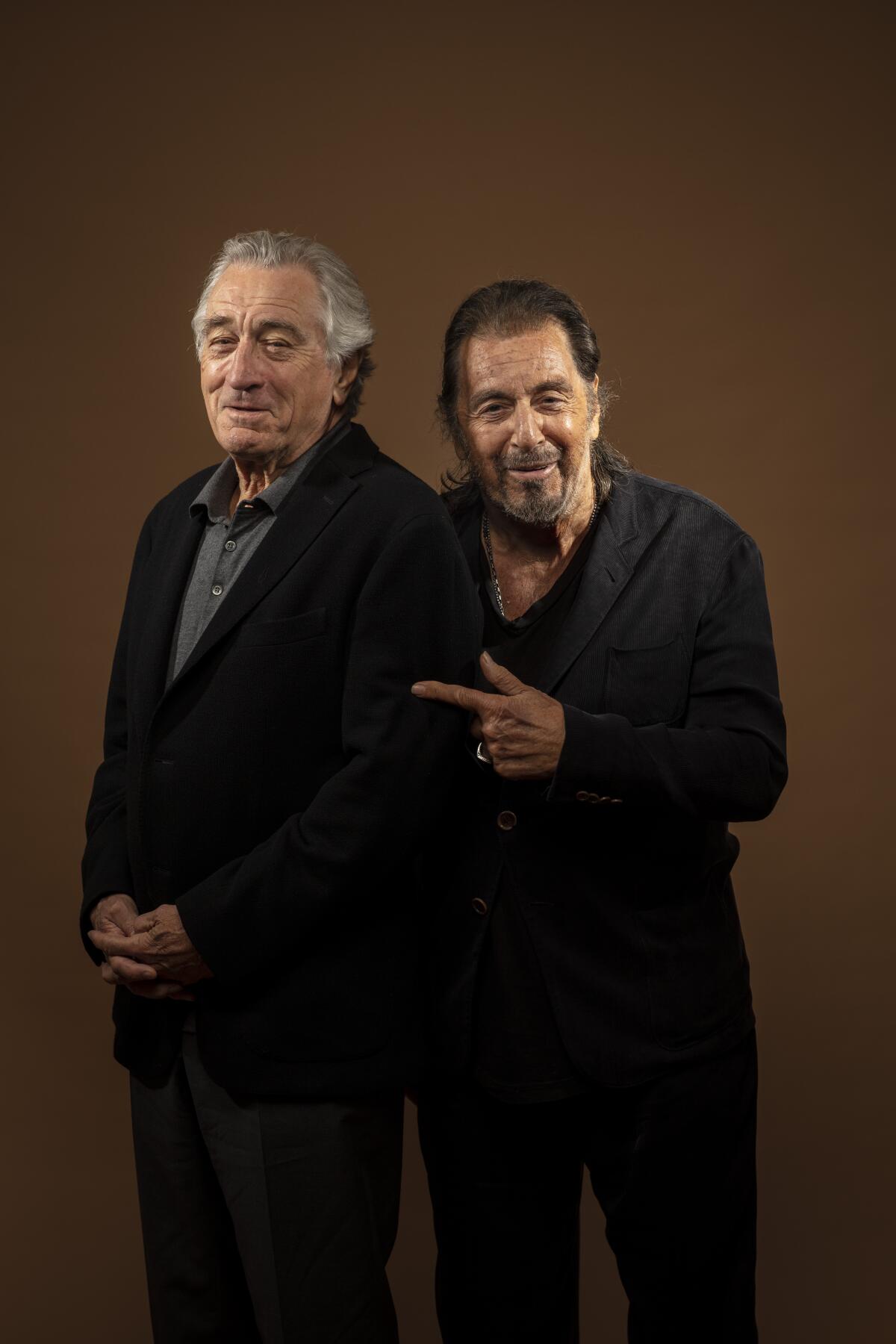 Robert De Niro wears a black blazer and stands next to Al Pacino who is dressed in black smiling, pointing to De Niro.