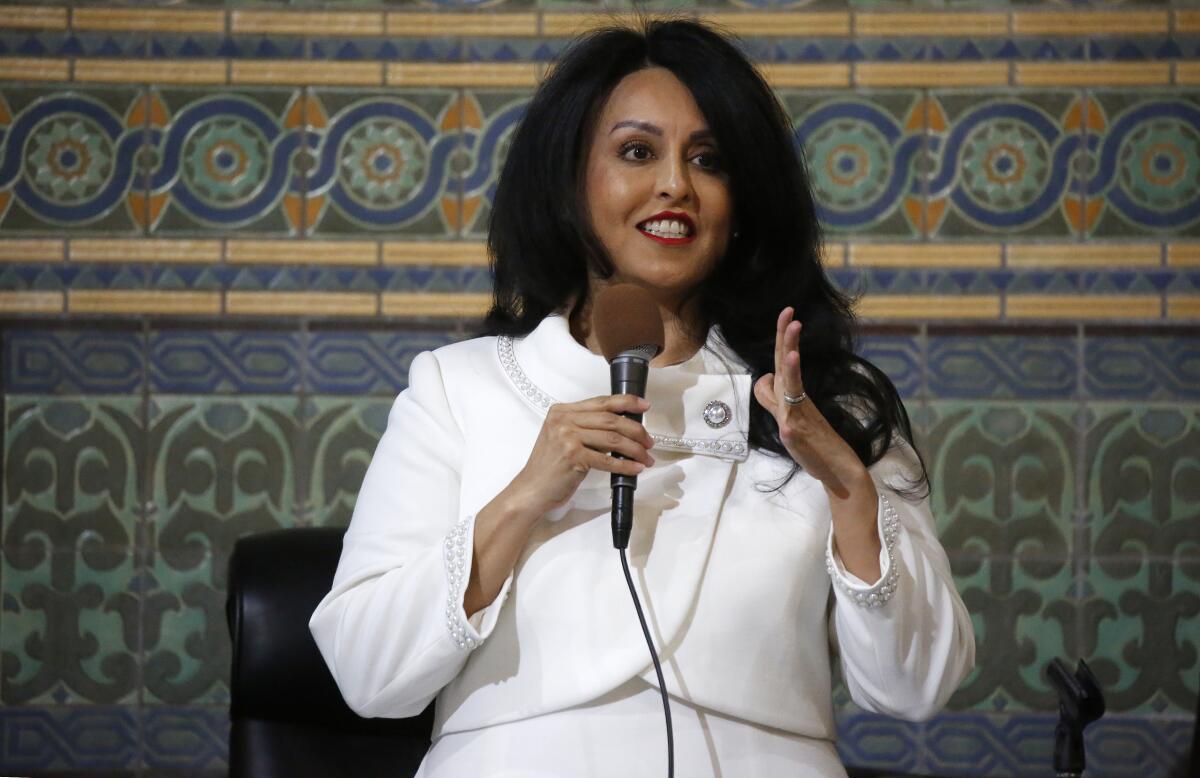 A woman with long black hair wearing a dressy white suit stands in front of tilework, holding a microphone