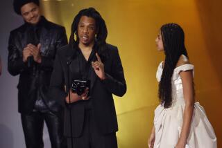 Jay-Z with dreads in a black suit standing on a stage next to Blue Ivy in a white dress.