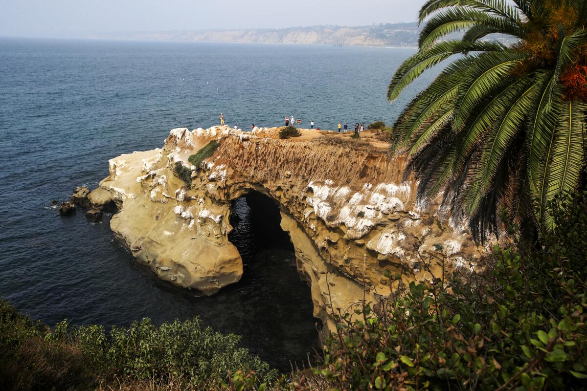 Seen from a distance, people stand atop land that juts out into the ocean, forming a cave beneath.