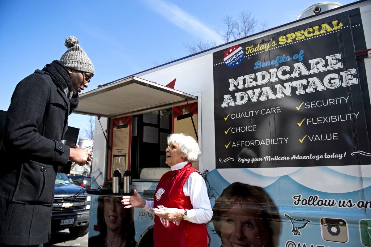 Two people stand in front of a trailer with a sign about Medicare Advantage plans.