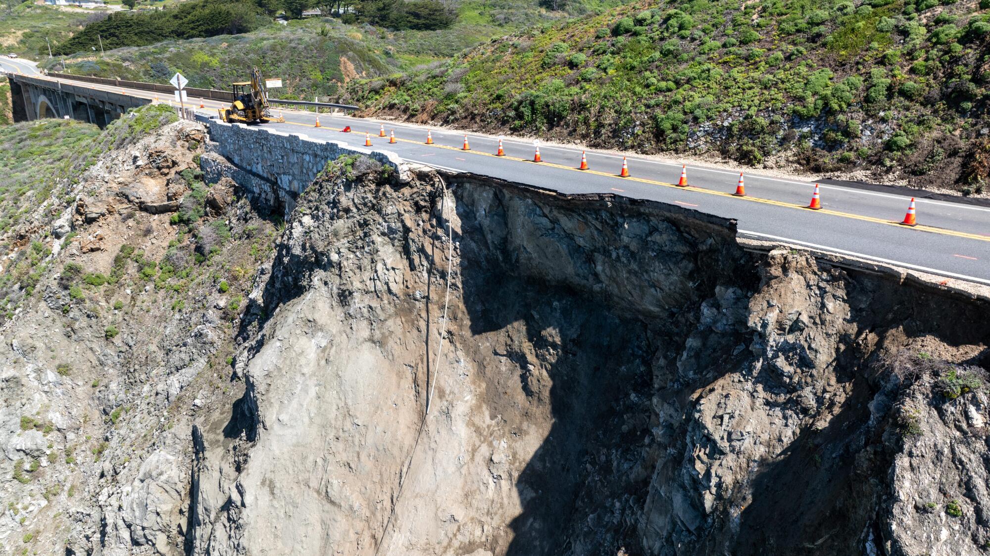 A line of orange cones sit in the middle of a damaged road on a cliff.