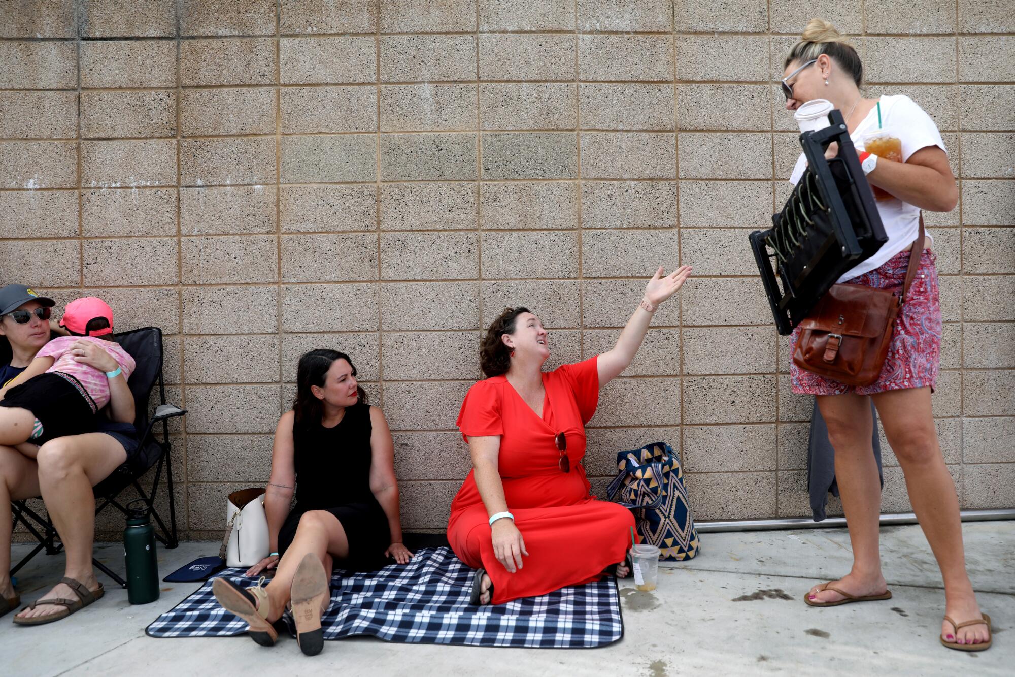 Katie Porter sits against a wall on a checkered blanket with a woman and gestures up to another woman standing next to them