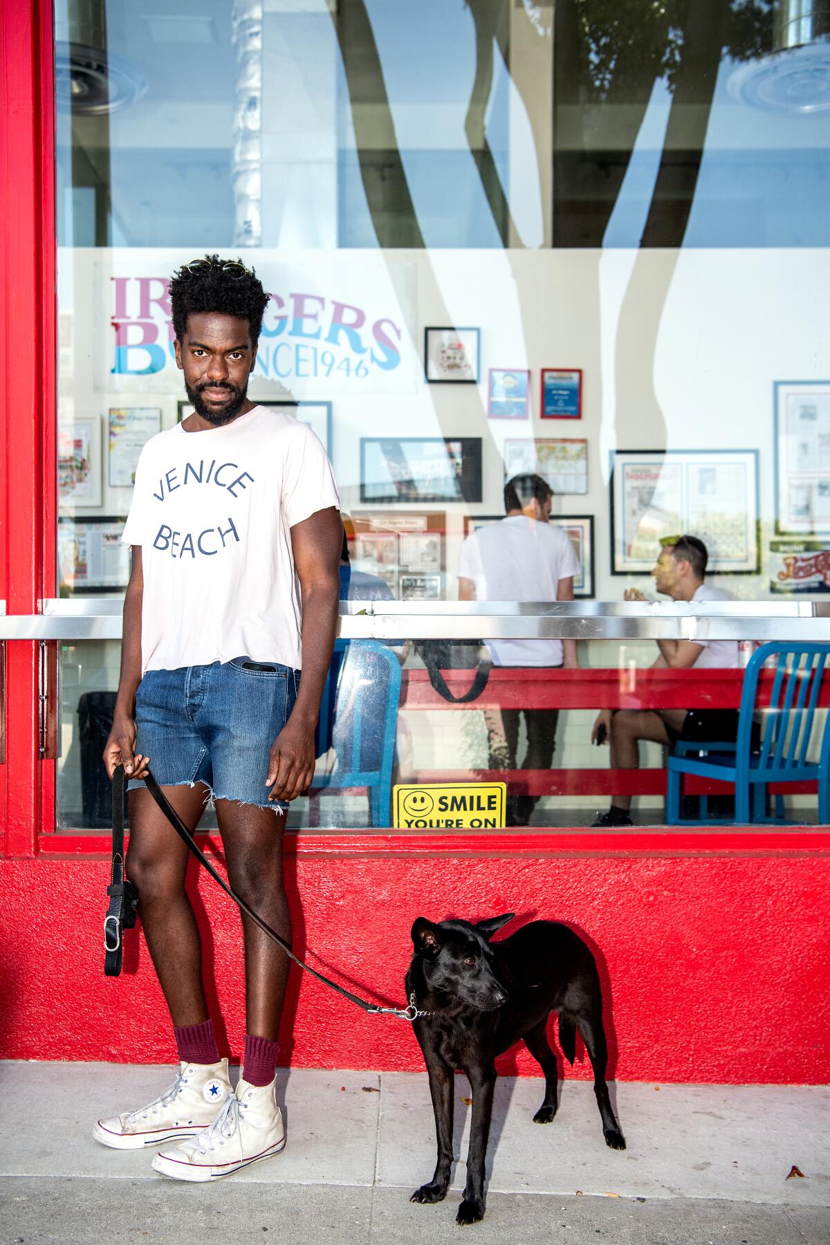 A photo of a man clad in denim shorts and a white shirt waiting in line outside of Irv's Burgers with his black dog, Mia.