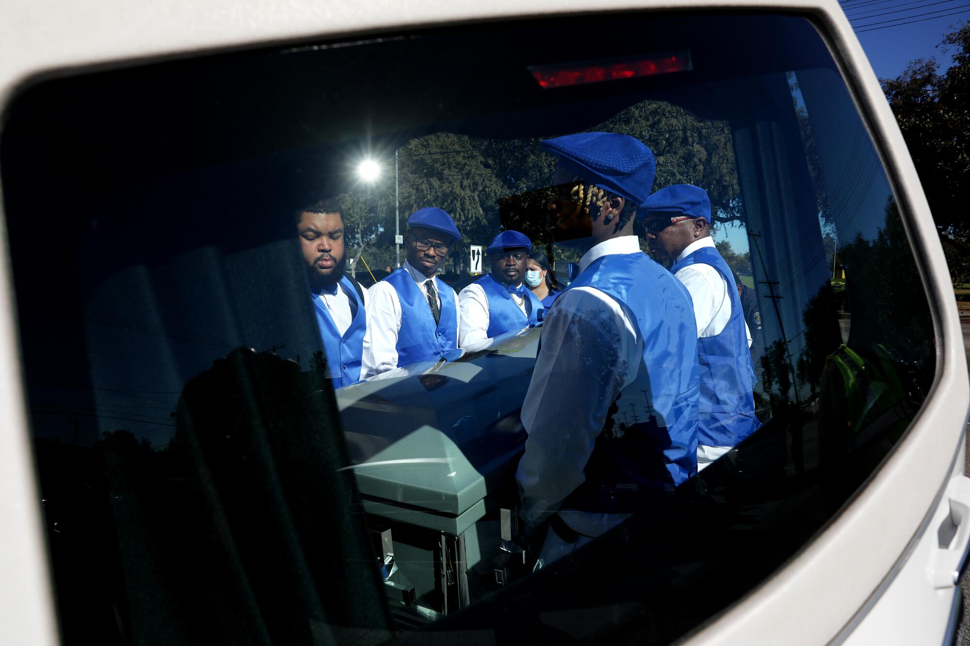 Pallbearers carrying a casket are reflected on a car door window 