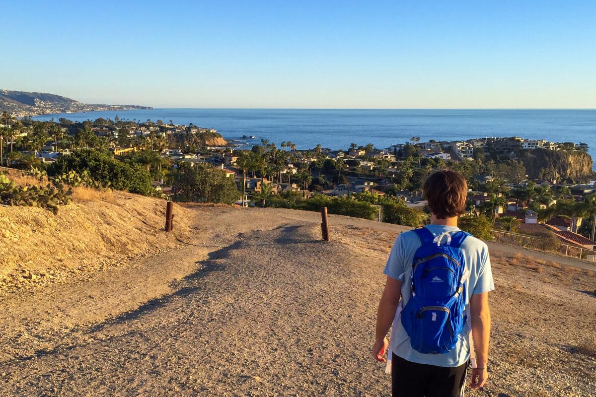 A man with a blue backpack walks on the trail with the ocean in the distance  
