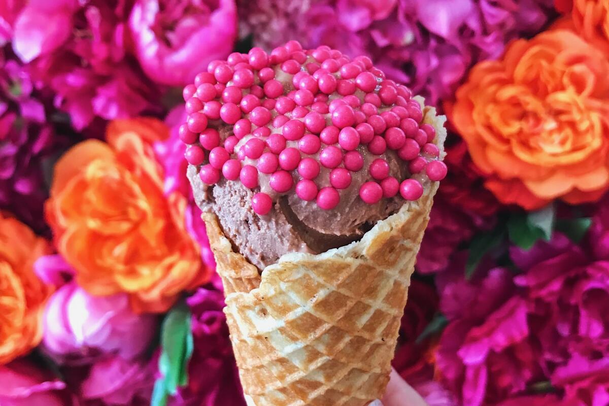 A hand holds a chocolate ice cream cone topped with strawberry chocolate orbs in front of a flower wall at Cafe de la Plage