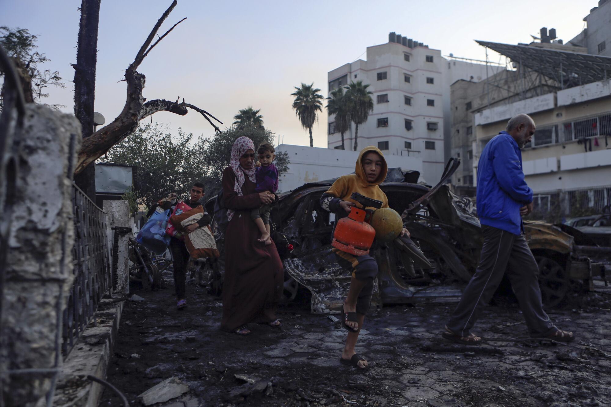 People carrying belonging walk amid the charred wreckage of a vehicle near multistory buildings 