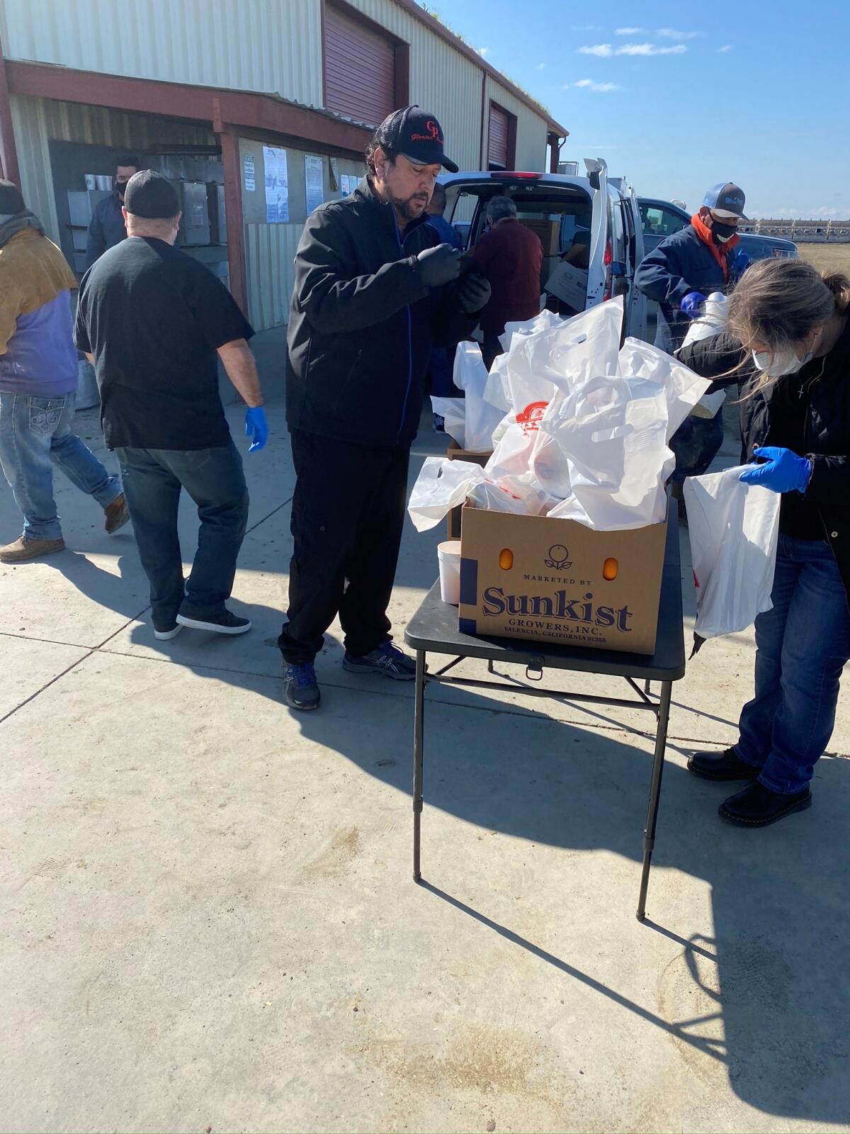 People handle bags of food outdoors on a folding table near a vehicle with an open cargo area.