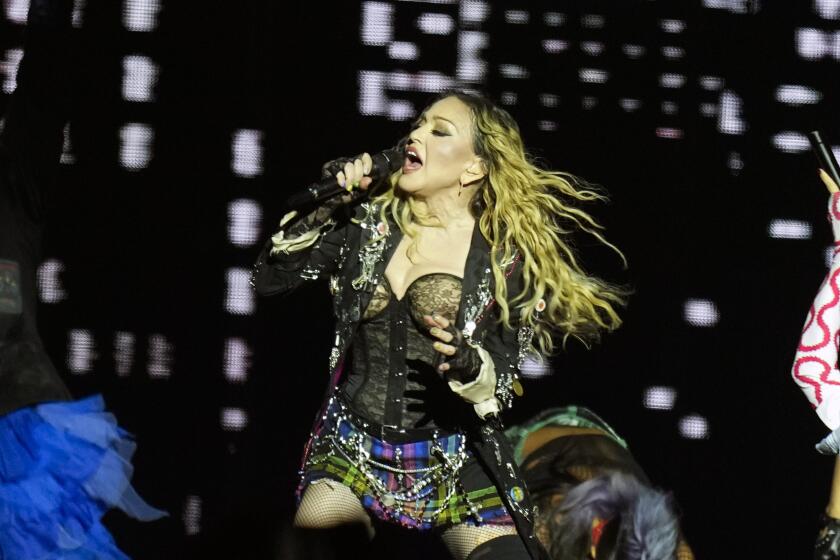 Madonna in a black corset, plaid mini skirt singing into a microphone in her right hand on a stage
