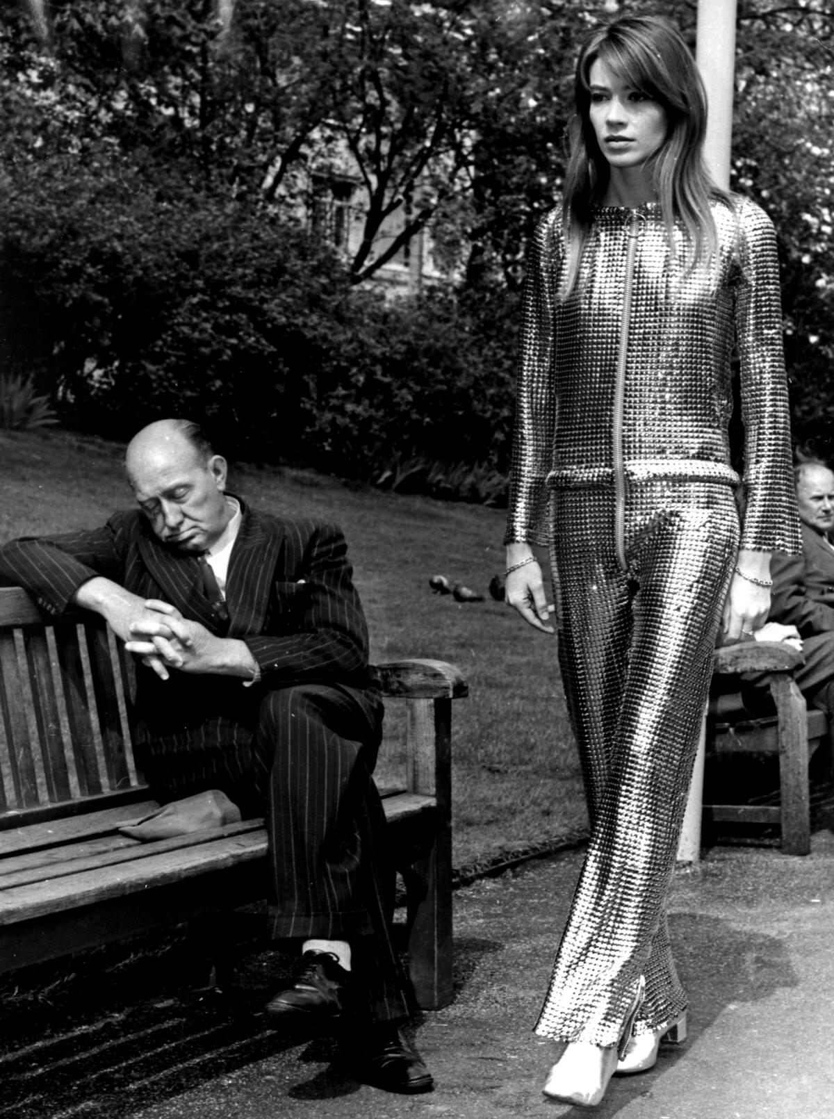 Françoise Hardy walks past a middle-aged man dressed in a suit and pretending to sleep on a park bench. 