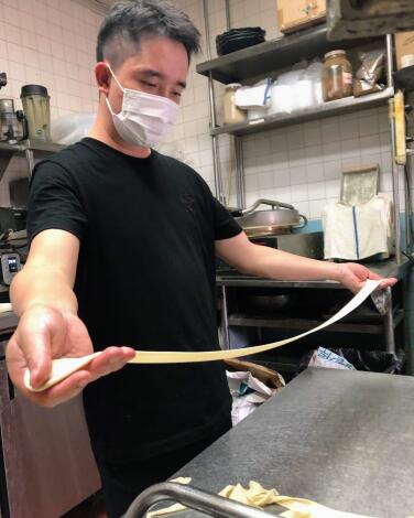A man in a restaurant kitchen stretches noodle dough, his arms outstretched