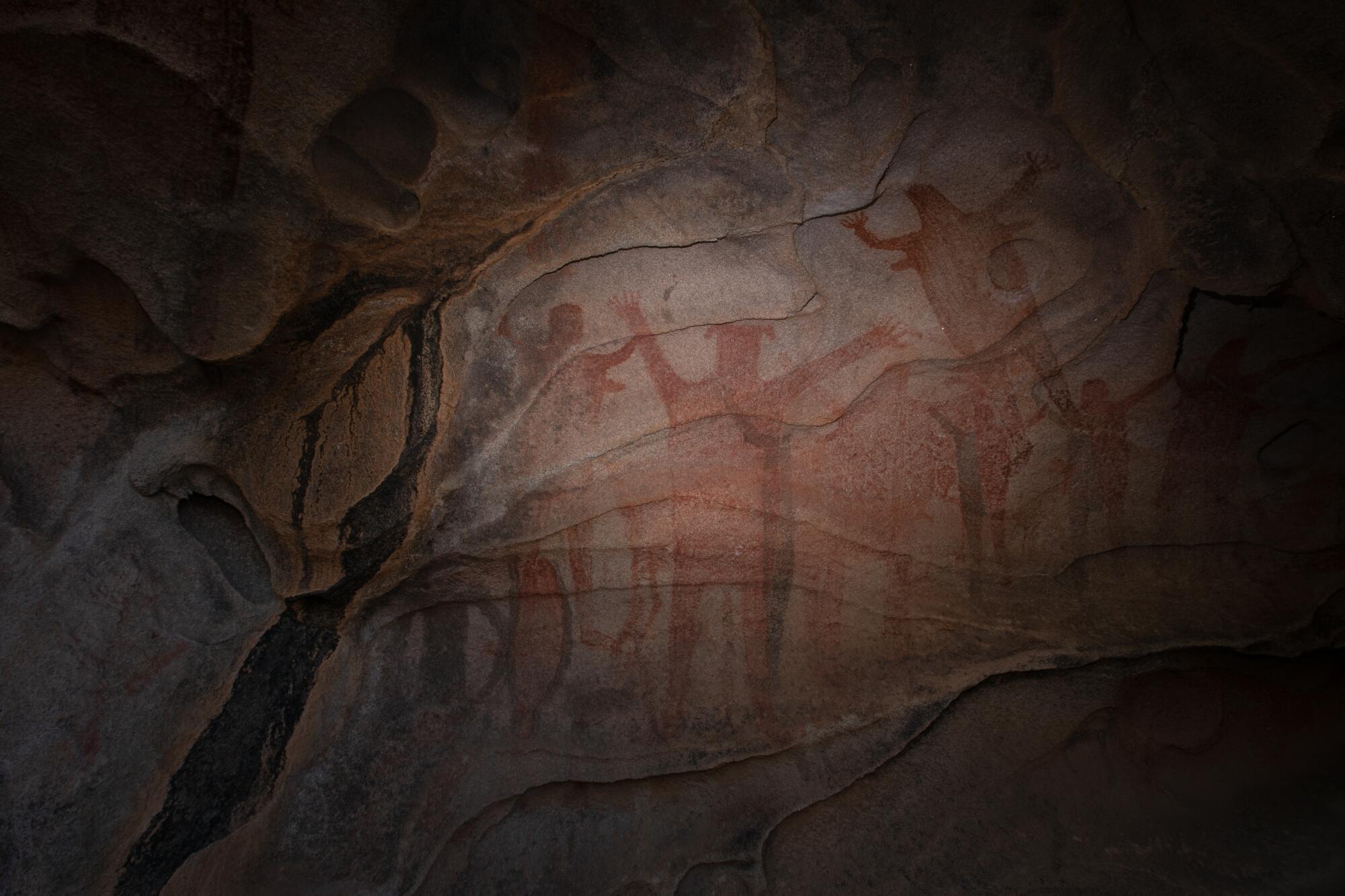 A cave wall, lit only by flashlight, showing ancient paintings of red humanoid figures.
