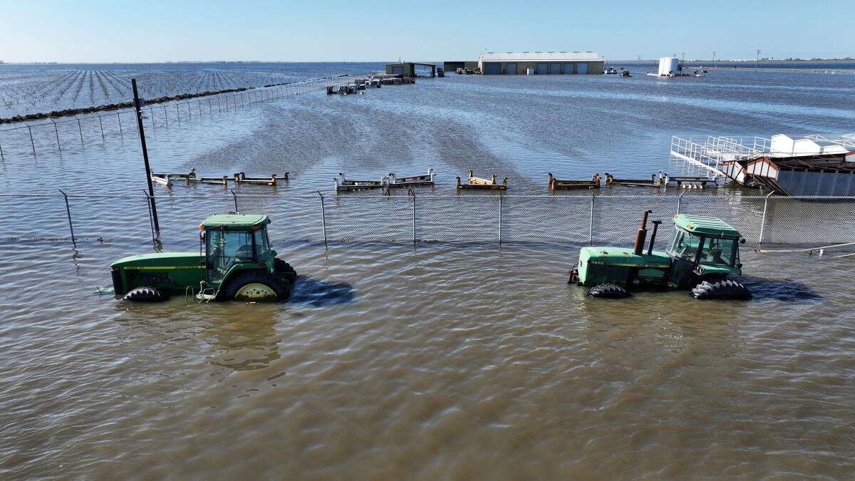 A pair of green farm tractors stand half submerged in muddy floodwaters.