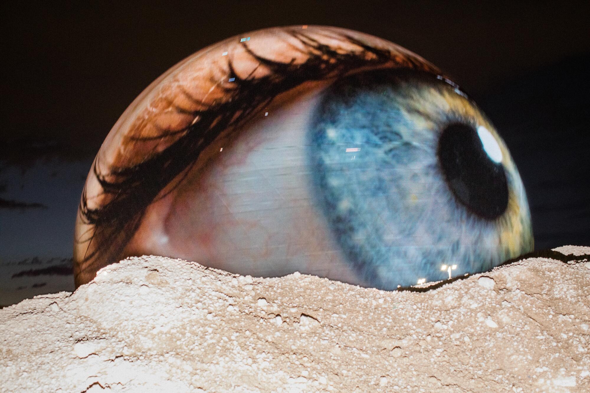 A spherical building covered in screens features a projection of an eyeball that seems to be emerging from the land