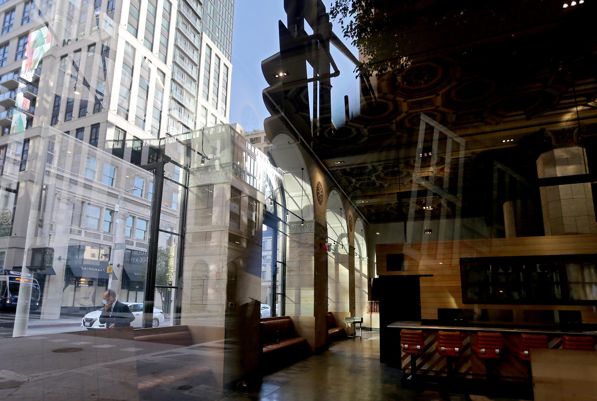 A man adjusting his mask is mixed into a jumble of reflections on the window of a closed restaurant in downtown L.A.