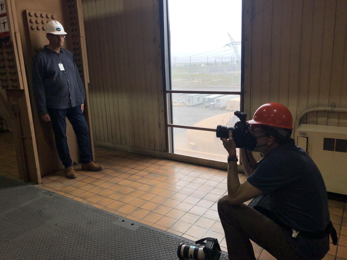 A photographer kneels while taking a photo of a man in a hard hat, who stands against an opposite wall.