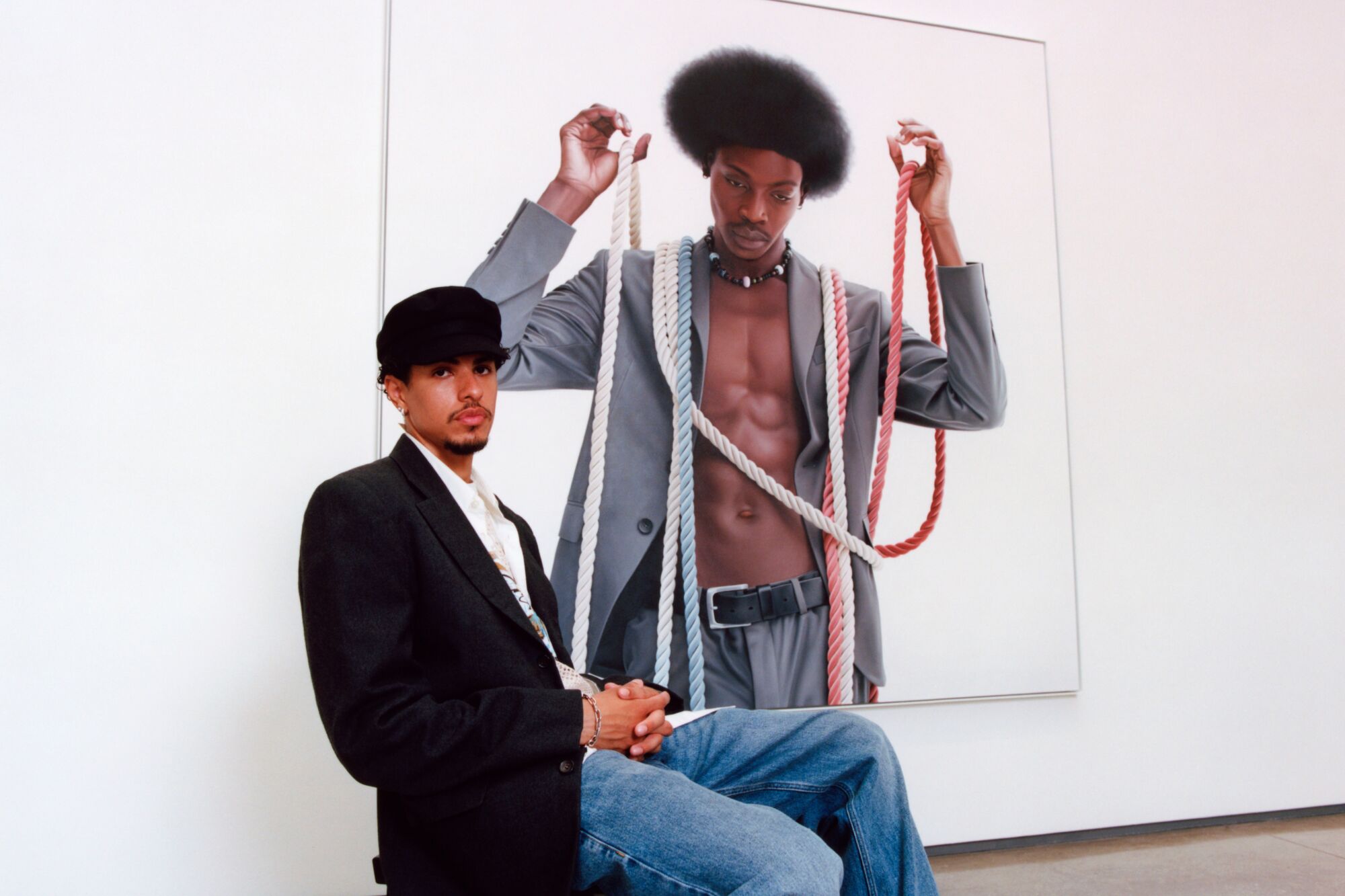Artist Delfin Finley sits on a stool in front of his painting of a man holding ropes in his hands.