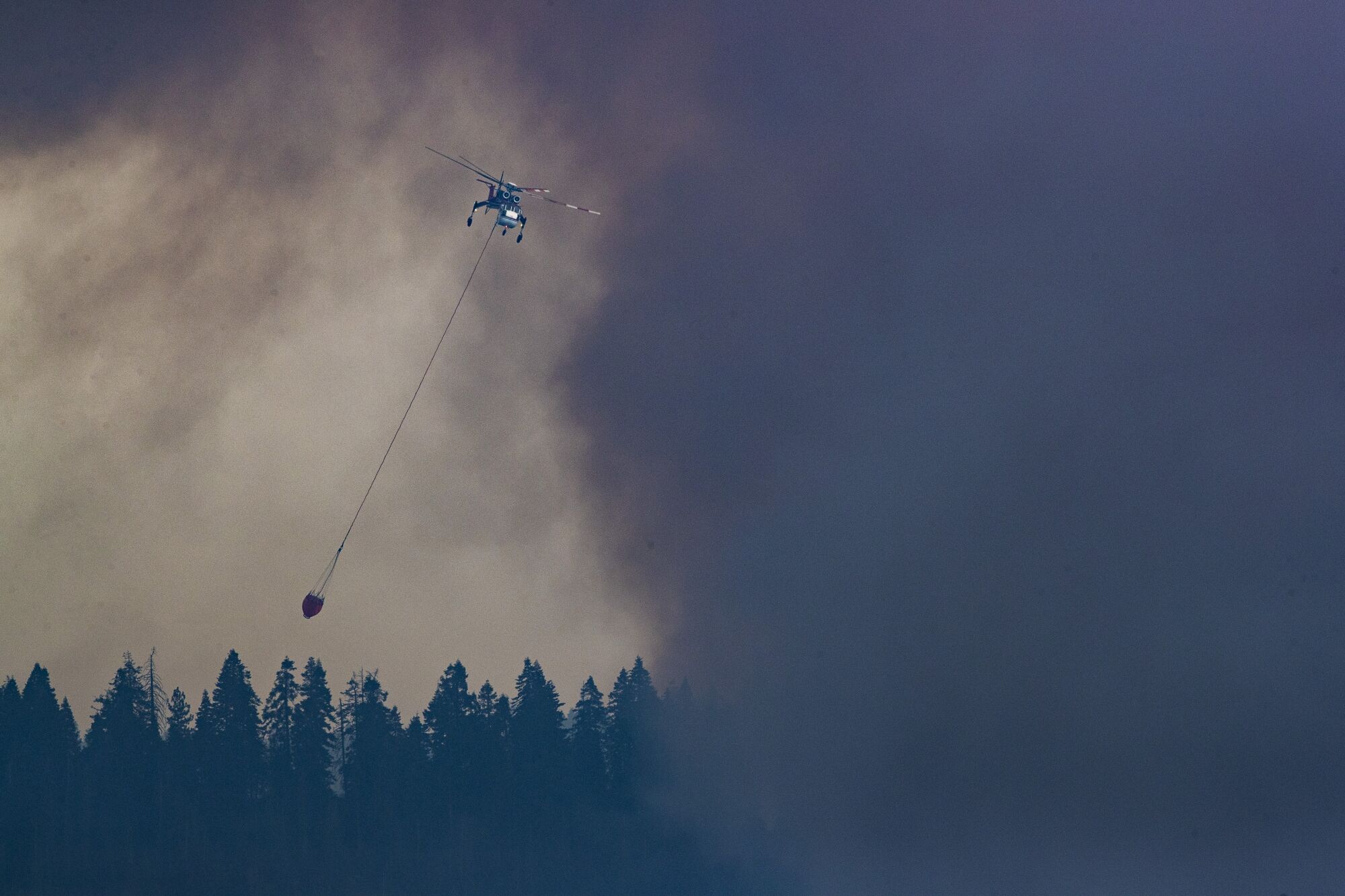 A water bucket dangles from a helicopter against a smoke-filled sky