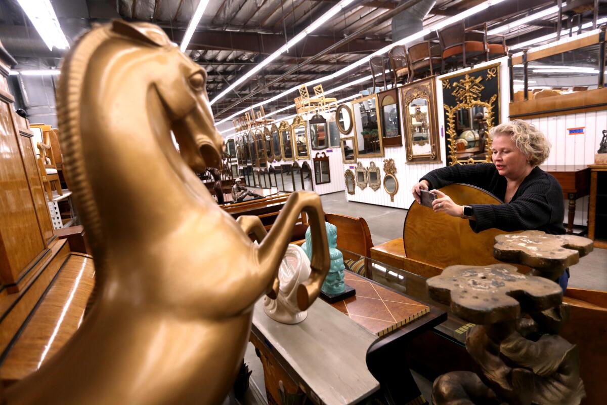 A woman with short blond hair takes a photo of a jade bust inside a giant warehouse full of furniture.
