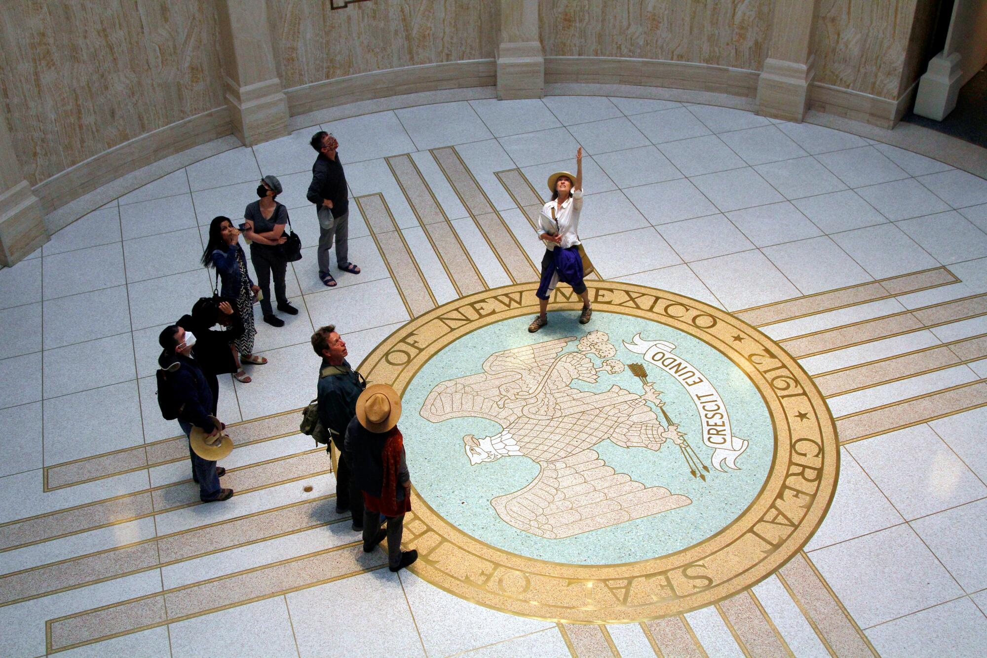 A bird's eye view of people looking up while standing in a round room, with a seal of New Mexico on the floor